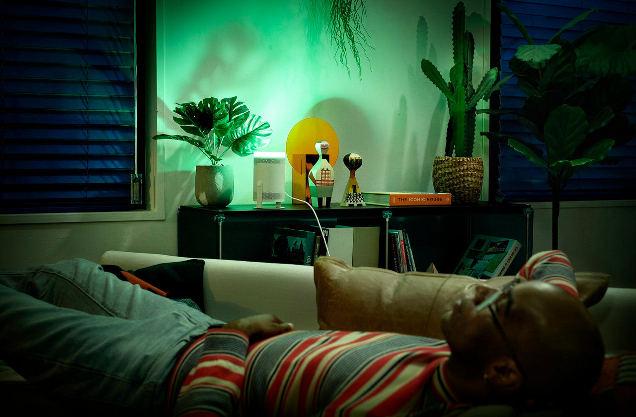 A man wearing a red, gray, and white striped long-sleeve shirt lies relaxed on a couch in a dimly lit living room with green ambient lighting. A bookshelf with plants and decor is visible in the background.