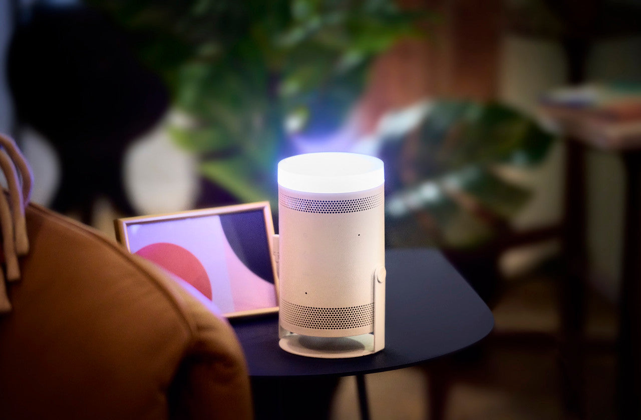 A modern cylindrical smart speaker or lamp in a warm off-white finish sits on a dark side table in a cozy living room setting, with a framed print and a brown leather chair blurred in the background.